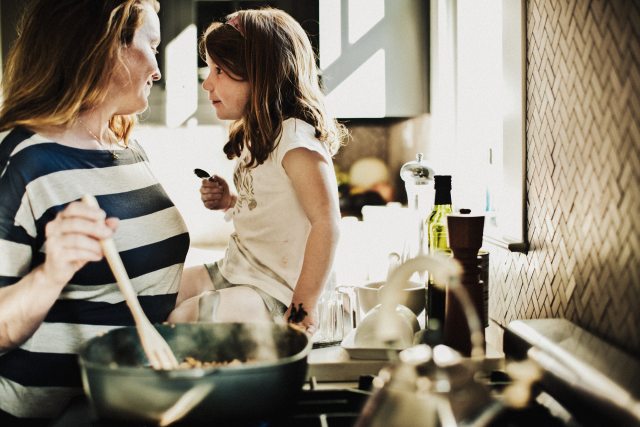 White woman stirring a pot on kitchen stove while smiling at a white child sitting next to her on the counter.